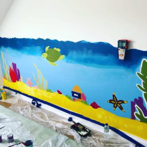 A wall in Sahara Care home painted blue with sea creatures and seaweeds, beneath an emergency alarm with a sheet and painting supplies on the floor