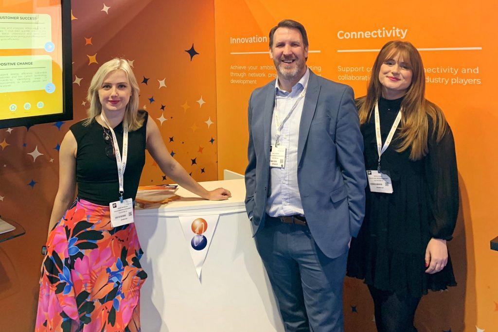 Orange background is stand at trade show, three people stand around a small white cabinet, smiling at the camera